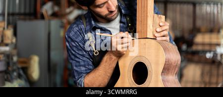 Primo piano di abili falegnami che marchiano il corpo della chitarra in legno con una matita in officina, indossando grembiule e camicia casual, mostrando attenzione, precisione e artigianato Foto Stock