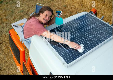 Donna felice che pulisce i pannelli solari sul tetto di un camper con una bottiglia spray e un panno. Foto Stock