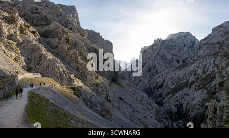 Un paesaggio di montagna panoramico con escursionisti su un sentiero tortuoso, circondato da ripide scogliere rocciose e un cielo luminoso dall'escursione Ruta del Cares in Spagna Foto Stock