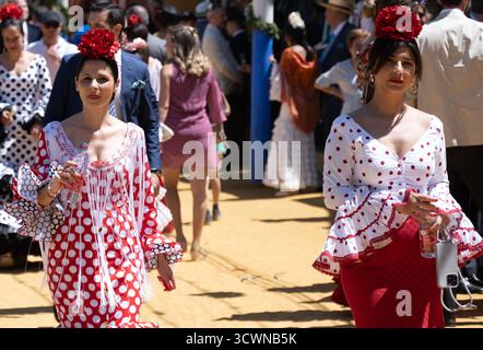 Siviglia, Spagna - 23 apr 23: Due donne in abiti tradizionali di flamenco con pois che camminano in un ambiente festoso all'aperto Foto Stock