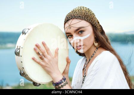 Bella donna sciamana con il viso che dipinge e rituale tamburello vicino al fiume all'aperto Foto Stock