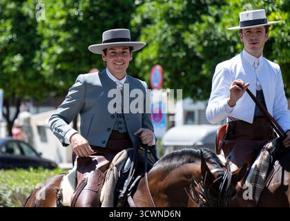 Siviglia, Spagna - 23 apr 23: Due uomini a cavallo indossano abiti e cappelli tradizionali, sorridenti, all'aperto in un ambiente soleggiato Foto Stock