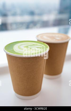 Due tazze di carta da asporto con latte matcha e cappuccino con latte art vicino alla finestra luminosa del bar. Primo piano. Profondità di campo ridotta Foto Stock