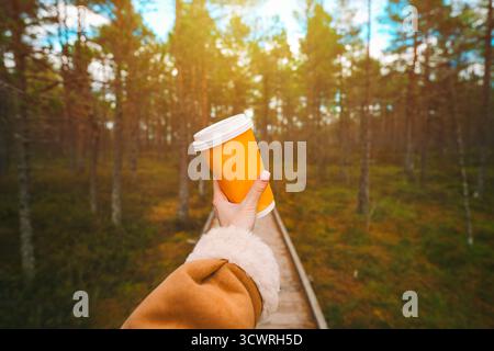 Tenere a mano una tazza di caffè da asporto sullo sfondo della foresta autunnale del parco al tramonto. Foto Stock