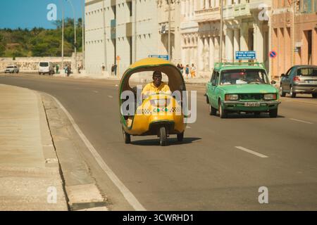 Havana, Cuba 2024 Apr16 - iconico taxi giallo cubano di cocco che guida lungo la strada sul Malecon in una giornata di sole. L'architettura coloniale si affaccia sul lungomare Foto Stock