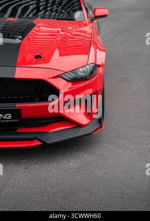 Red Ford Mustang su una strada. Angolo anteriore sinistro della pony car americana in una giornata nuvolosa, che si riflette in una carrozzeria lucente. Foto Stock