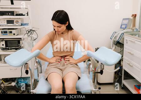 Una giovane donna siede su una sedia ginecologica tenendo l'addome in dolore. Il fastidio addominale durante la consultazione segnala possibili problemi ginecologici. W Foto Stock