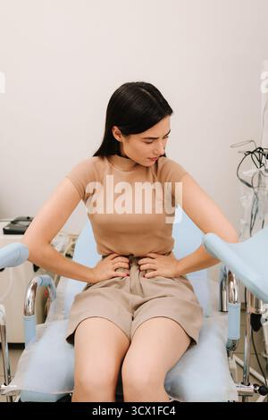 Una giovane donna siede su una sedia ginecologica tenendo l'addome in dolore. Il fastidio addominale durante la consultazione segnala possibili problemi ginecologici. W Foto Stock