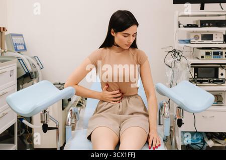 Una giovane donna siede su una sedia ginecologica tenendo l'addome in dolore. Il fastidio addominale durante la consultazione segnala possibili problemi ginecologici. W Foto Stock