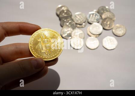 Moneta Golden Bitcoin tenuta in mano contro un tavolo con varie monete d'argento, a simboleggiare lo scontro tra valute digitali e fiat. Foto Stock