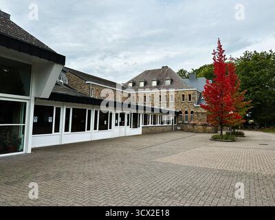 Banneux, Belgio - 20 ottobre 2023: Un tranquillo cortile belga accanto a un edificio in pietra con vetrine e un albero rosso brillante. Santuario di o Foto Stock