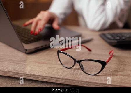 Vista ravvicinata degli occhiali sulla scrivania in legno con computer portatile e calcolatrice come un lavoratore digita, che illustra l'ausilio visivo, il lavoro sullo schermo e la produttività dell'ufficio Foto Stock