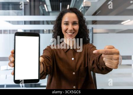 Giovane donna d'affari sorridente, con in mano uno smartphone con uno schermo bianco vuoto e che lo indica, mostrando la tecnologia in un ambiente d'ufficio moderno Foto Stock