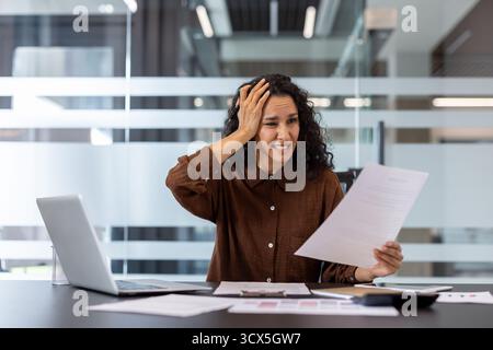 Donna d'affari stressata che tiene un documento e stringe la testa, che soffre frustrazione e ansia per problemi di lavoro e per un lavoro burocratico travolgente nel suo armadio Foto Stock
