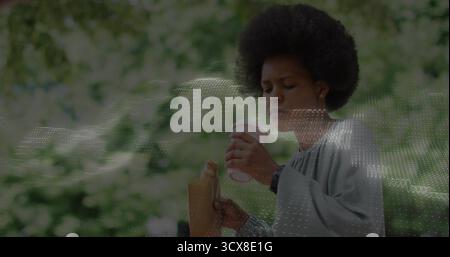 Tenendo la tazza di caffè bianco vicino alla bocca, donna in camicetta grigia in parco con sacchetto di carta Foto Stock