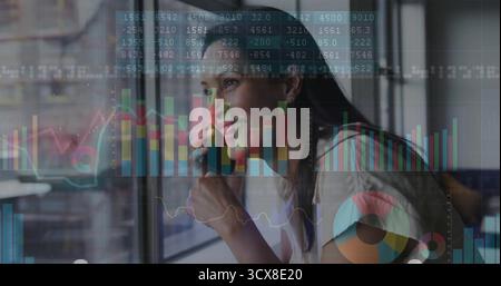 Donna sorridente che indossa un blazer che tiene lo smartphone all'orecchio alla finestra dell'ufficio, con grafici finanziari Foto Stock