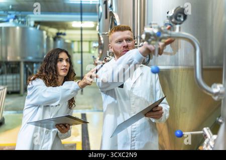 Gli ingegneri chimici e un tecnico del controllo qualità stanno controllando i serbatoi e le valvole di fermentazione in acciaio inox in una moderna fabbrica di birra, garantendo la conformità Foto Stock