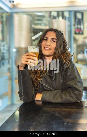 Donna sorridente con i capelli ricci che indossa una giacca in pelle, che tiene in mano un bicchiere di birra chiara mentre si rilassa in un bar della birreria, con serbatoi industriali visibili Foto Stock