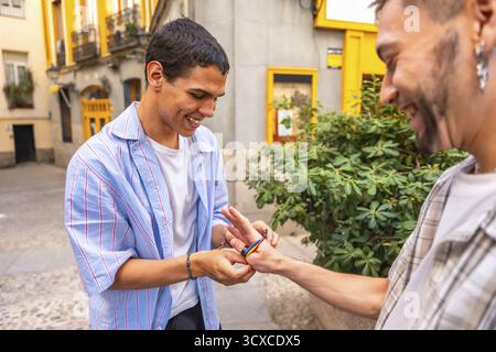 Il giovane gay lega un braccialetto arcobaleno al polso del suo partner, sorridendo in una strada urbana che celebra orgoglio, amore, accettazione e gioiosa scommessa di connessione Foto Stock