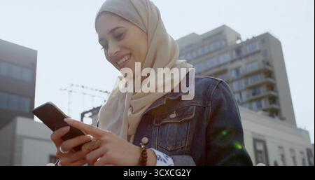 Donna sorridente che indossa sciarpa e giacca in denim che controlla lo smartphone in City plaza con anello e bracciale Foto Stock