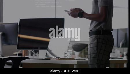 Lavoratore in ufficio in piedi che indossa una camicia grigia che controlla lo smartphone alla scrivania, con monitor e spazio di copia Foto Stock