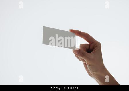 Una mano che regge delicatamente un cartoncino bianco bianco su uno sfondo bianco pulito con una luce naturale soffusa. La carta può rappresentare l'attività, il branding o Foto Stock