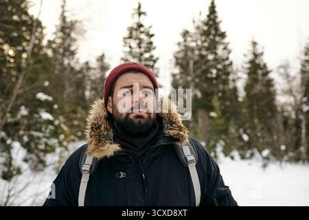 Ritratto di un uomo barbuto adulto con giacca calda e cappello in piedi con zaino in legno invernale che guarda la macchina fotografica Foto Stock