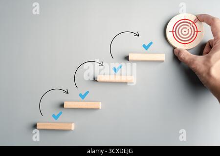 Elenco di controllo degli obiettivi e passaggio verso il successo con blocchi di legno disposti come scale, segni di spunta e bersaglio Foto Stock