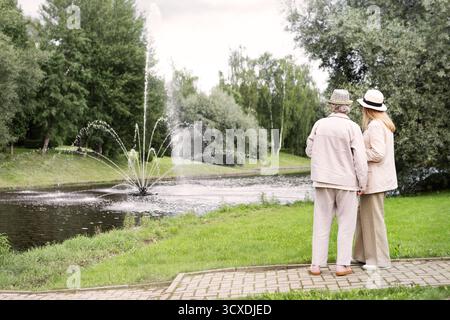 Due eleganti donne anziane che viaggiano insieme, in piedi accanto alla fontana del parco, indossano abiti leggeri e cappelli, si godono una conversazione tranquilla e rilassante Foto Stock