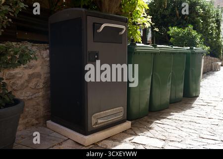 Un contenitore per rifiuti comuni alto, grigio scuro, si trova su un percorso in pietra accanto a una linea di contenitori di riciclaggio verdi standard Foto Stock