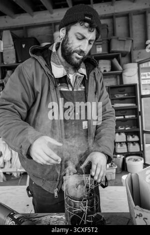 Un apicoltore prepara un fumatore di alveare all'interno di un laboratorio di legno presso la Dancing Bee Farm, catturato in bianco e nero per enfatizzare l'artigianato, il fumo e la consistenza Foto Stock