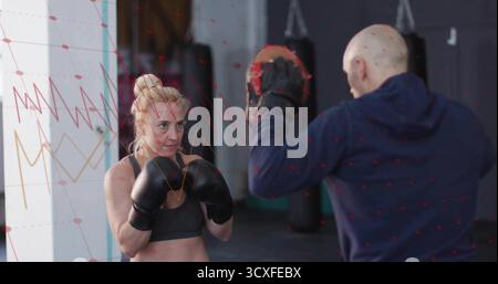Pugile da allenamento indossando un reggiseno sportivo che colpisce i guanti a fuoco nella palestra di pugilato, con guanti e borse da pugno Foto Stock