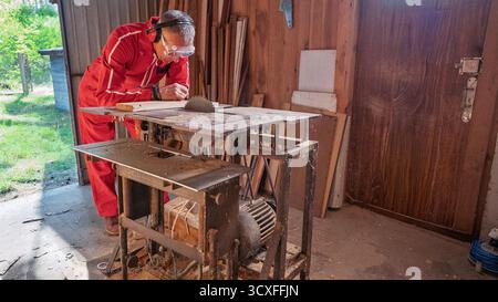 Un uomo studia un pezzo di legno appoggiandosi su un banco da lavoro con una sega circolare. Foto Stock
