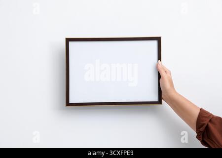 Donna che appende una cornice vuota per diplomi su un muro bianco, primo piano. Mockup per il design Foto Stock