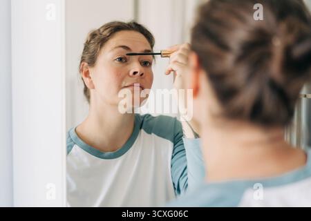 Una donna caucasica di quarant'anni applica il mascara agli occhi mentre guarda allo specchio. Foto Stock