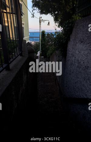 Scala in uno stretto passaggio tra edifici e pareti su una scogliera al tramonto con una sezione di una fabbrica sullo sfondo Foto Stock