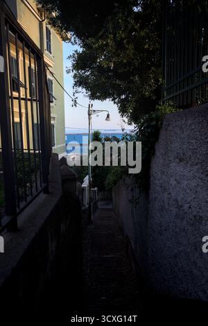Scala in uno stretto passaggio tra edifici e pareti su una scogliera al tramonto con una sezione di una fabbrica sullo sfondo Foto Stock