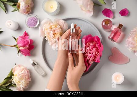 Mani femminili con bottiglie di olio essenziale, candele, sale marino e bellissimi fiori di peonia su sfondo chiaro Foto Stock