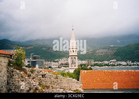La storica torre della chiesa si innalza sopra le vecchie mura di pietra della città vecchia di Budva, con edifici moderni e montagne verdi nebbiose sullo sfondo di un film Foto Stock
