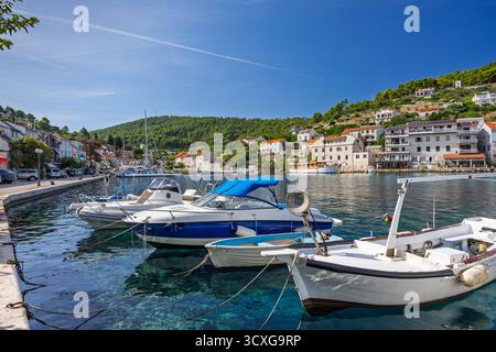Un luminoso porto con barche ancorate lungo una banchina di pietra, case pastello e bianche su una collina, e un cielo azzurro limpido, che trasmette un mare tranquillo Foto Stock