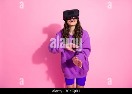 Giovane donna in un abbigliamento sportivo alla moda che utilizza il visore per la realtà virtuale su un vivace sfondo rosa Foto Stock
