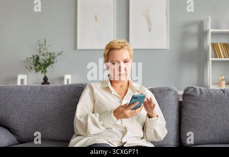 Donna matura con capelli biondi corti seduta su un comodo divano, leggendo e-mail di lavoro sullo smartphone. Foto Stock