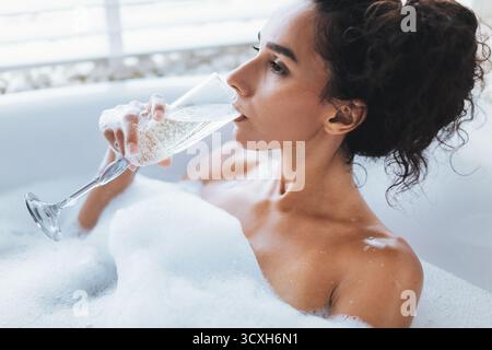 Rilassati in un bagno di bolle mentre sorseggi un bicchiere di bevanda frizzante in serata Foto Stock