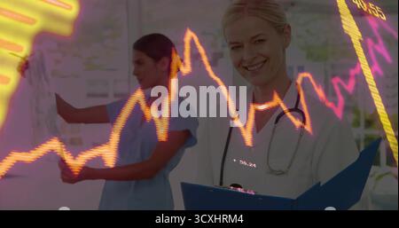 Medico sorridente che tiene in camera gli appunti e lo stetoscopio, con un collega che scrive sulla lavagna Foto Stock