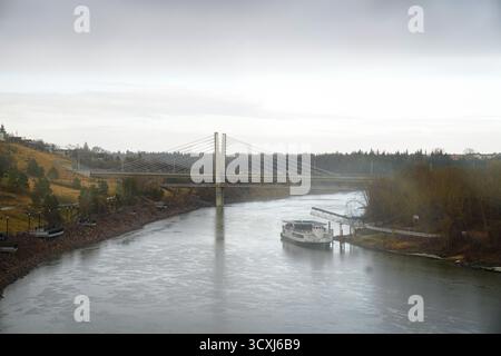 Barca ormeggiata lungo il fiume North Saskatchewan vicino a un ponte moderno e alla passerella sul fiume, evidenziando un equilibrio tranquillo Foto Stock