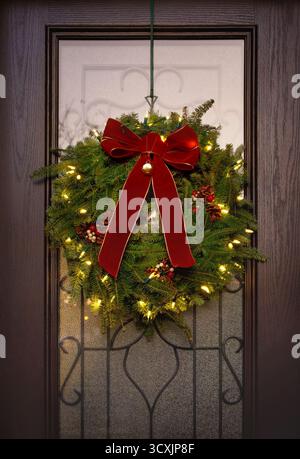 Tradizionale corona di Natale Balsam Fir appesa su una porta d'ingresso. Fiocco rosso e luci natalizie. Foto Stock