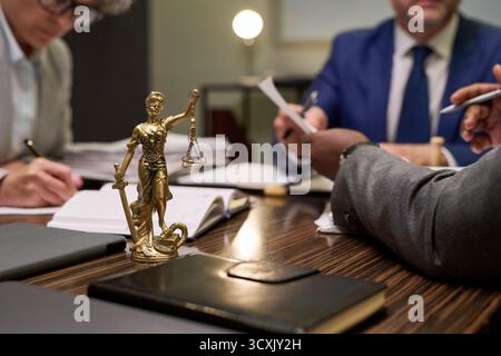 Primo piano della statua di Lady Justice in piedi sul tavolo con un gruppo multietnico di uomini di mezza età che discutono documenti legali in ufficio, mani che tengono penne e documenti, si concentrano sul tema della legge Foto Stock