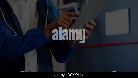 Tenendo in mano il tablet, il tecnico indossa una giacca blu che tocca il touch screen in officina, insegna a strisce rosse Foto Stock