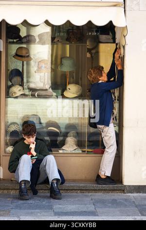 Un giovane uomo si siede premurosamente vicino a un negozio di cappelli, mentre una donna più anziana adatta la tenda in una strada tranquilla. Foto Stock