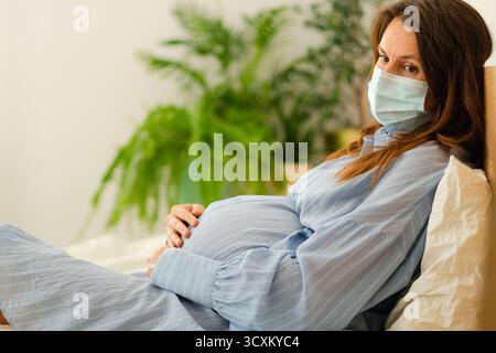 La mamma indosserà una maschera di sicurezza mentre si riprende a letto. Importanti misure di controllo delle infezioni durante il periodo di gravidanza sensibile. Foto Stock
